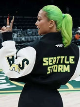 A woman with bright neon green hair poses on a basketball court wearing a black and white Seattle Storm varsity jacket with 'SEATTLE STORM' printed in neon green on the back.