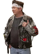 A man wearing a heavily embellished black leather jacket with studs, a white 'DUDE' headband, a dark gray t-shirt, and light blue jeans, standing in a kitchen setting.