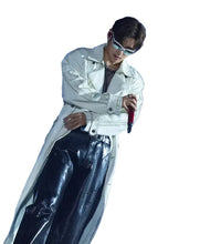 A male performer on stage wearing a long white leather-like coat, shiny black pants, and reflective sunglasses while holding a microphone against a blue bokeh-lit background.