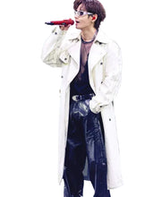A male singer performing on stage wearing a long cream-colored trench coat over a sheer black top and black pants, holding a red microphone.