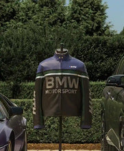 A BMW Motorsport motorcycle jacket displayed on a mannequin outdoors, with hedges and cars visible in the background. The jacket is dark gray with a blue upper section, white lettering, and a small red, white, and blue Kith logo on the chest.
