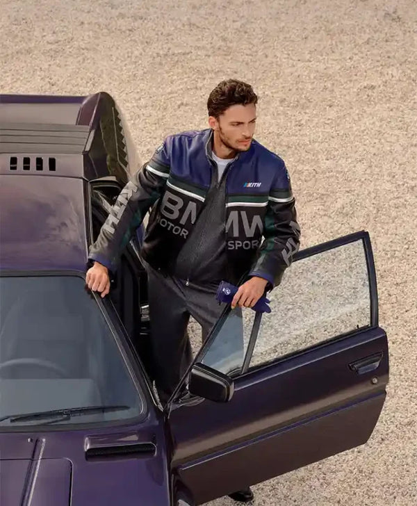 A man wearing a BMW Motorsport leather jacket and matching trousers stands beside an open door of a dark purple car on a gravel surface.