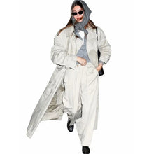A woman modeling an oversized light beige trench coat with a matching oversized pair of wide-leg trousers, styled with a gray hooded scarf, gray top, black sunglasses, and black shoes against a plain white background.