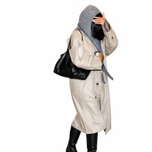 A person wearing a long light beige trench coat over a gray hoodie, with black knee-high boots and a black shoulder bag, posed against a plain white background.