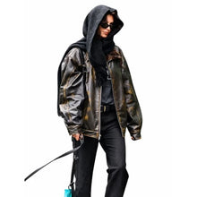 A person wearing an oversized distressed black leather jacket with gold-toned burnishing, a black hoodie, black pants, and sunglasses, holding a black strap against a plain white background.