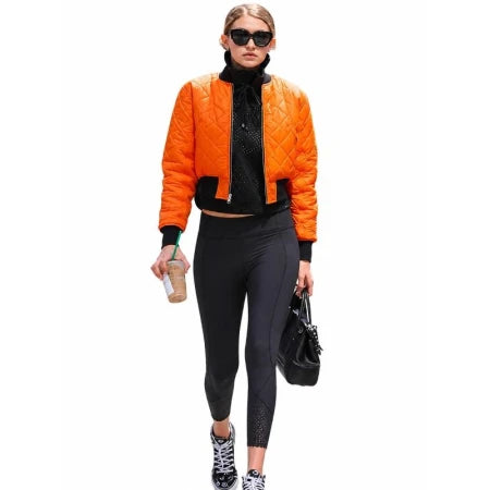 A woman modeling a bright orange quilted bomber jacket over a black turtleneck, paired with black leggings, black-and-white sneakers, sunglasses, and a black handbag, against a plain white background.