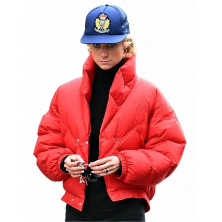 A model wearing a bright red cropped puffer jacket over a black turtleneck, paired with a blue baseball cap featuring a crest logo, set against a plain white background.