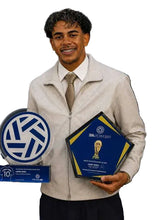 A smiling man in a light beige zip-up jacket, white shirt, and beige tie holds two award plaques in front of a dark blue sponsor backdrop. The left plaque is clear with a blue and white Volkswagen logo, and the right plaque is dark blue with gold and white details featuring the FIFA World Cup trophy design.