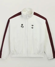 A white zip-up track jacket with dark red shoulder stripes and black embroidered graphics on the chest, displayed against a plain light background.