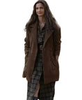 A woman wearing a brown double-breasted coat over a dark plaid shirt and skirt, standing outdoors with a blurred vehicle in the background.