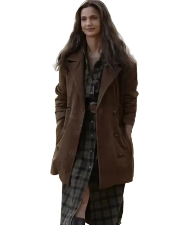 A woman wearing a brown double-breasted coat over a dark plaid shirt and skirt, standing outdoors with a blurred vehicle in the background.