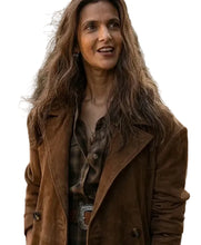 A woman with long, wavy brown hair is wearing a brown suede-like jacket over a dark plaid shirt, standing outdoors in front of a blurred background of trees and a fence.
