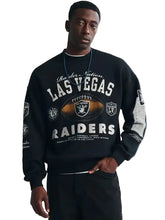 A man wearing a black Raiders Nation Las Vegas Raiders crewneck sweatshirt with white and gold graphics, paired with black pants, against a light gray studio background.