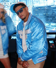 A man wearing a shiny light blue satin varsity jacket with white stripes, a white t-shirt, khaki pants, and a light blue cap, standing in a modern indoor setting with screens in the background.
