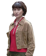 A woman standing outdoors in front of police cars, wearing a tan cropped jacket over a red top and olive green pants.