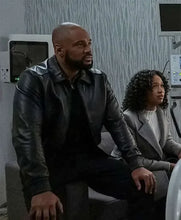 A man in a black leather jacket and a woman in a gray blazer sitting in a room with medical or technical equipment in the background.