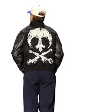 A person shown from behind wearing a black bomber-style jacket with a large distressed white skull and crossbones graphic on the back, navy pants, and a white baseball cap with a purple brim, standing against a dark stage-like background.