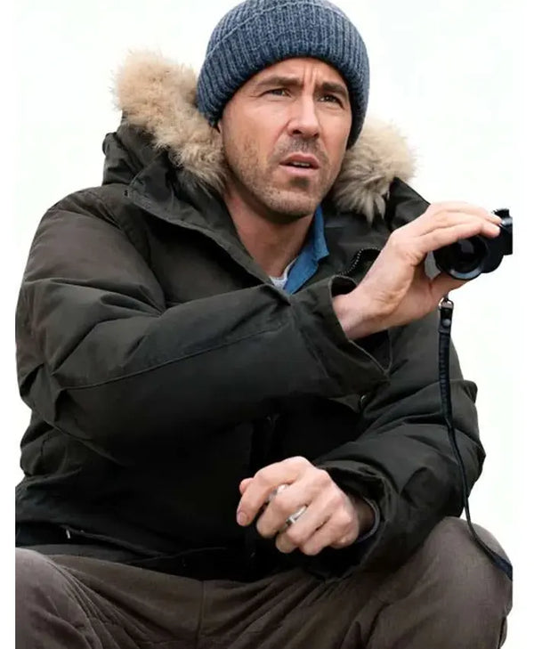 A man wearing a dark winter parka with a fur-trimmed hood, a blue knit beanie, and brown pants, holding a small camera against a plain white background.