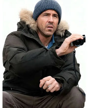A man wearing a dark winter parka with a fur-trimmed hood, a blue knit beanie, and brown pants, holding a small camera against a plain white background.