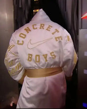A person shown from behind wearing a white hooded jacket with gold lettering that reads 'CONCRETE BOYS' and a Nike swoosh, along with a matching gold belt.