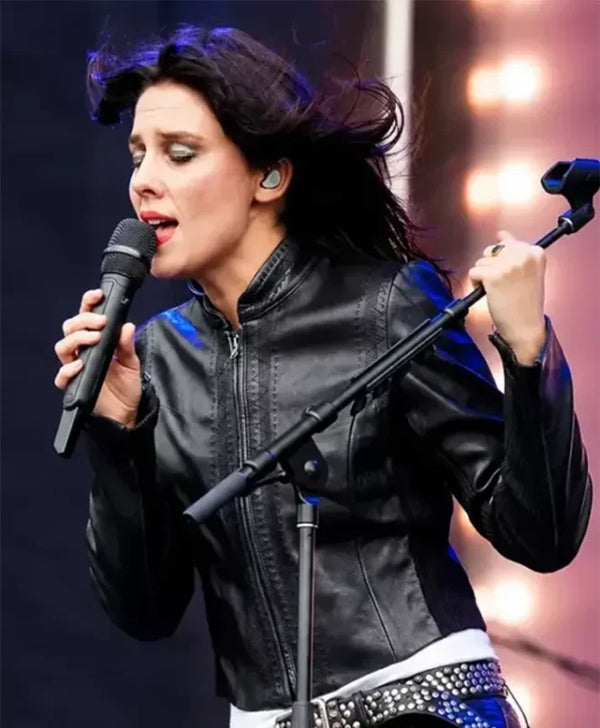 A female singer performing live on stage, wearing a black leather jacket and holding a microphone, with a blue and white studded belt visible.