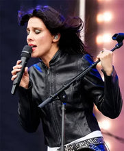 A female singer performing live on stage, wearing a black leather jacket and holding a microphone, with a blue and white studded belt visible.