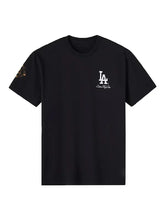 A black short-sleeve crewneck t-shirt featuring a white 'LA Champions' logo on the left chest and a small gold geometric patch on the right sleeve, displayed against a solid white background.