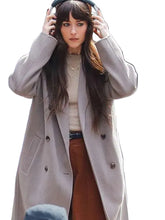 A woman wearing a long, double-breasted light beige coat over a cream sweater and brown pants, holding over-ear headphones on a city street.