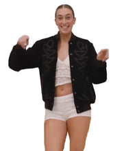 A woman smiling and posing in a black varsity-style bomber jacket with white lace crop top and white shorts in an indoor studio setting.
