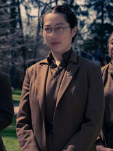 A woman wearing round glasses, a brown blazer over a dark top, and a small lapel pin, standing outdoors with trees in the background.