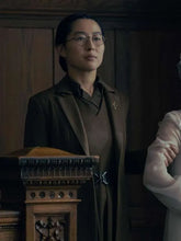 A woman wearing a dark brown tailored blazer over a matching brown top, with round wire-frame glasses, standing indoors near a carved wooden lectern. Part of another person in a pale dress is visible at the right edge.