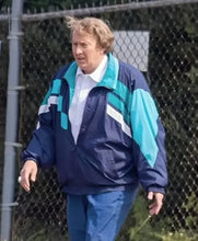 A man walking outdoors in front of a chain-link fence, wearing a navy blue and turquoise colorblock windbreaker jacket over a white collared shirt and blue pants.