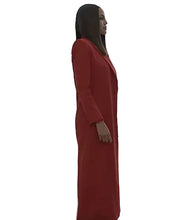 A woman wearing a long, red, tailored dress or coat stands in profile inside a modern glass-walled office with a city skyline visible outside.
