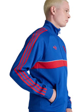 A man in profile wearing a blue Adidas track jacket with red stripes and a red chest stripe, photographed against a light gray studio background.