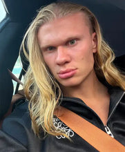A close-up selfie of a man with long blonde hair wearing a black zip-up hoodie and a seatbelt inside a car.