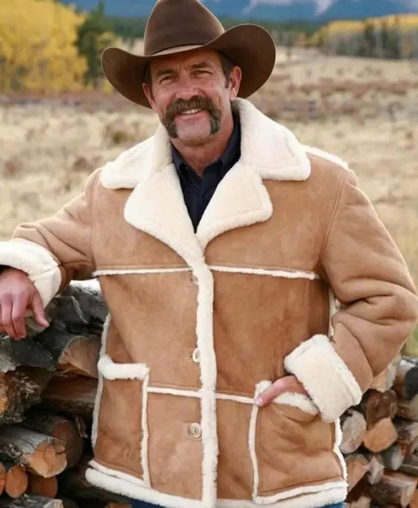 A smiling man outdoors wearing a tan shearling-style jacket with cream lining, a brown cowboy hat, and standing beside a stack of firewood.