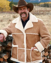 A smiling man outdoors wearing a tan shearling-style jacket with cream lining, a brown cowboy hat, and standing beside a stack of firewood.