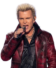 A close-up shot of a male singer with platinum blonde hair, wearing a dark red leather jacket over a black shirt, holding a microphone on stage with a blue lit background.