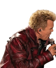 A man with short blond hair wearing a dark red leather jacket and holding a microphone, shown in profile against a blurred stage background.