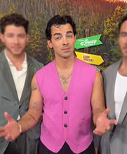A man wearing a bright pink sleeveless vest stands in front of a Disney Camp Camp backdrop, with two other men partially visible on either side.