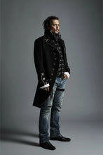 A man wearing a long black military-style coat with silver buttons and trim, a black bow tie, a patterned vest, and light blue jeans, posed against a dark gray studio background.