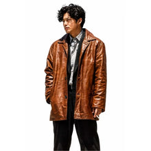 A man wearing a brown leather jacket over a gray shirt and black pants, standing against a plain white background.