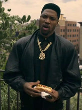 A man wearing a black leather jacket, black shirt, and a large gold chain necklace with a pendant, holding a hot dog outdoors with buildings and greenery in the background.