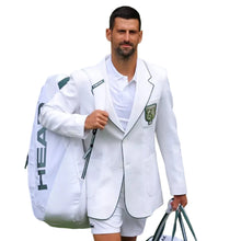 Novak Djokovic walking on a green grass court wearing a white blazer with green piping, white shirt, and white shorts, carrying a large white HEAD tennis bag.
