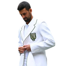 A man in a white blazer with green piping and a Wimbledon crest patch on the chest, standing in a stadium with a blurred crowd in the background.