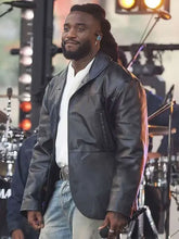A man on a stage wearing a black leather jacket over a white shirt and light blue jeans, with band equipment visible in the background.