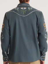 A man viewed from the back wearing a dark slate blue western-style shirt jacket with white contrast stitching, a central eye motif, and gold-toned sleeve embroidery.