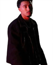 A man wearing a black jacket with silver studs and a black shirt, photographed against a plain white background with dramatic red lighting.