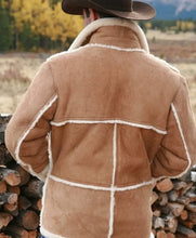 A man viewed from behind wearing a tan shearling-style jacket with white trim and a brown cowboy hat, standing outdoors in front of stacked firewood with a blurred field and mountains in the background.