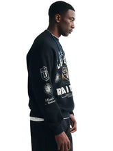 A man in profile wearing a black graphic sweatshirt with white and gold lettering and patches, paired with black pants, against a plain light background.